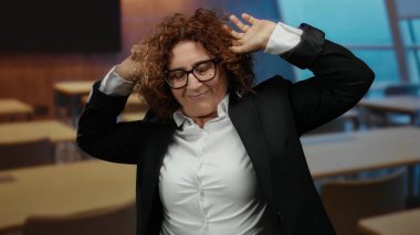 Ahşap masalı ve modern tasarımlı, gözlüklü ve takım elbiseli orta yaşlı esmer bir kadın, bir sınıfta uyukluyor ve el kol hareketi yapıyor..