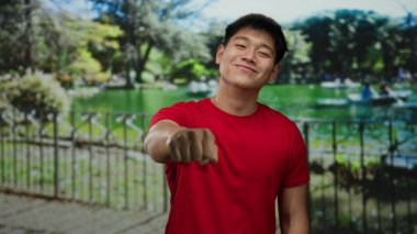 Kırmızı gömlekli genç adam yeşil ağaçlar ve dingin göl arka planıyla güneşli bir park ortamında yumruğunu uzatarak açık havada dostça bir hava yaratıyor..