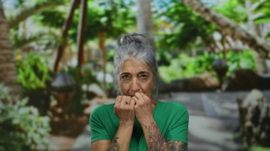 Yeşil tişörtlü, gri saçlı ve dövmeli yaşlı bir kadın açık hava ortamında kaldırılmış ellerle korkusunu dile getirerek yemyeşil bir parkta ürkmüş görünüyor..