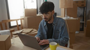 Yeni evde dizüstü bilgisayarda çalışan genç bir adam güneşli bir dairede paketlenmemiş kutularla çevrili, sıradan bir yaşam tarzını gösteriyor..