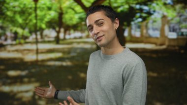 Gri kazaklı genç Hispanik adam sanki yapraklı bir park yolu sunar gibi iki elini uzatır;.