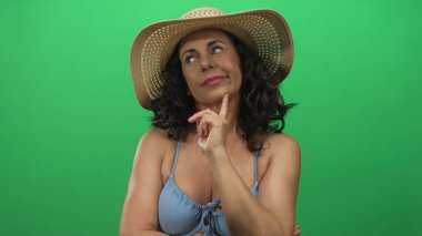 Orta yaşlı, bikinili ve güneş şapkalı bir kadın yeşil bir arka plan duvarına karşı durarak sakin bir yaz tatili havasını vurguluyor..