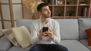 Genç adam, elinde telefonuyla koltukta oturuyor, arka planda kitaplık ile düşünceli bir şekilde bakıyor, rahat ve içten bir atmosferi öneriyor rahat bir oturma odasında..