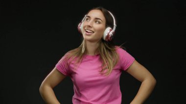 Pembe tişörtlü, kulaklıklı, izole edilmiş siyah arka planda müzik dinleyen mutlu bir gülümseme, kapalı gözler, kalçasında eller, neşe ifade eden bir kadın..