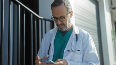 Tıbbi kıyafetli olgun İspanyol bir adam hastanenin önünde duruyor, telefonuna odaklanmış, profesyonel ve kentsel bir ambiyansı var..