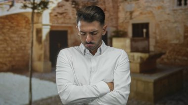 Bir adam, güneş ışığıyla aydınlatılmış dar bir sokakta, yıpranmış taş bir binanın dışında kolları bağlı duruyor, beyaz bir gömlek giyiyor ve ciddi bir ifadeyle reddediliyor;.