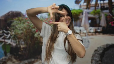 Güneşli bir sahilde gülümseyen bir kadın elleri parlak çiçekler ve açık mavi gökyüzü ile çevrili bir çerçeve hareketi yapıyor.