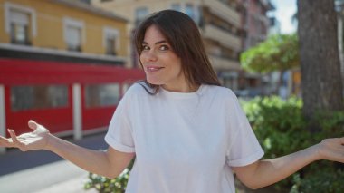 Gündelik beyaz gömlekli bir kadın arka planda yeşillik ve kırmızı binalar olan güneşli bir şehir sokağında kafa karışıklığı içinde açık havada şaşkın bir ifadeyi canlandırıyor..