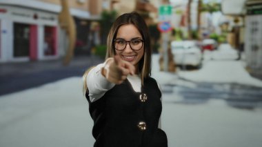 Kadın, enerjik bir şekilde, açık havada, gözlük ve siyah yelek takan, şehir sahnesine güven ve pozitiflik saçan, enerjik bir şekilde parmağıyla işaret ediyor..