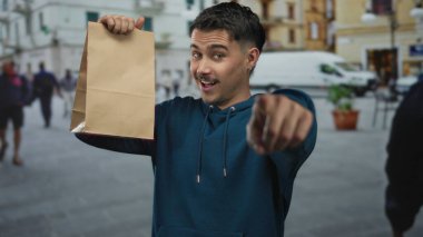 Sokak ortasında elinde kese kâğıdıyla gezen genç İspanyol adam gülümsüyor ve etrafı hareketli şehir manzaralarıyla çevrili..