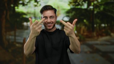 Parkta sakallı genç adam iki orta parmağı havada, etrafı yeşilliklerle çevrili, siyah gömlekli, dışarıda duygularını ifade eden kaba bir jest yapıyor..