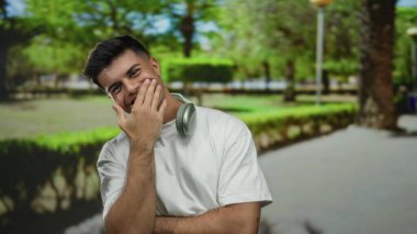 Kulaklıklı genç adam, yeşil ağaçlar ve patikalarla çevrili güneşli bir parkta gülümsüyor, rahat bir açık hava havasını yansıtıyor ve gündelik bir stili yansıtıyor..