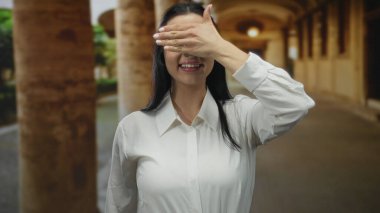 Beyaz tişörtlü genç İspanyol kadın eski bir üniversite avlusunda gülümserken eliyle gözlerini kapatıyor..