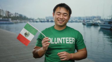 Deniz kenarında Meksika bayrağı taşıyan genç bir adam, yeşil bir gönüllü tişört giyiyor, limanda neşeli bir ifade sergiliyor..