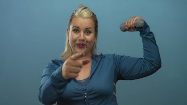 Genç sarışın kadın, mavi stüdyo duvarına karşı dambılla çıplak kolunu esnetiyor; güven motivasyonu.