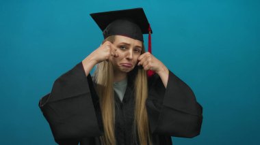 Mezuniyet kıyafetli genç kadın, mavi arka plana karşı neşeli bir ağlama hareketi yapıyor, kep ve cübbe giyiyor, yeni mezun olmuş biri gibi alaycı üzüntüsünü dile getiriyor..