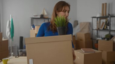 Mavi elbiseli kadın, yeni apartman dairesinde taşınan kutuları taşıyor. Etrafı tıka basa doldurulmuş eşyalarla çevrili ve elinde bitki tutarken gülümsüyor..