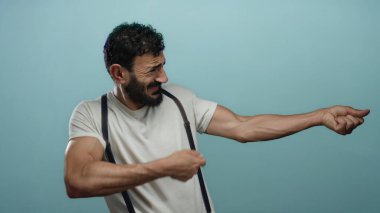 Sakallı İspanyol bir adam. Mavi arka planda ip çekiyormuş gibi yapıyor. Espri anlayışı ve kas gerilimi gösteriyor..