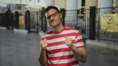 Çizgili tişörtlü genç adam başparmağını kaldırıyor ve kent geçmişiyle gülümsüyor. Şehir sokaklarında pozitif ve kendine güvenini gösteriyor..