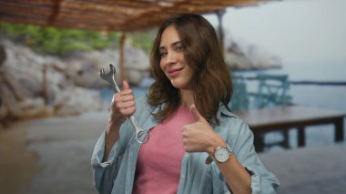 İspanyol kadın bir İngiliz anahtarı tutuyor, baş parmağıyla gülümsüyor, arka planda deniz olan bir sahilde duruyor, işi ve rahatlamayı harmanlıyor..