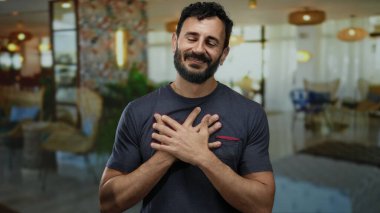 Otel lobisinde elleri göğsünde gülümseyen sakallı İspanyol bir adam, zarif dekorlu bir ev ortamında sıcaklığı ve dostluğu iletiyor..