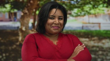 Kırmızı süveterli genç ve iri yarı Latin kadın yemyeşil ağaçlarla güçlendirilmiş güneşli bir parkta çapraz kollarıyla gülümsüyor..