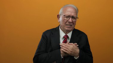 Takım elbiseli yaşlı işadamı canlı turuncu arka plana karşı göğüs ağrısı gösteriyor. Tecrit ortamında klasik yaşlı sağlık stresi gösteriyor..