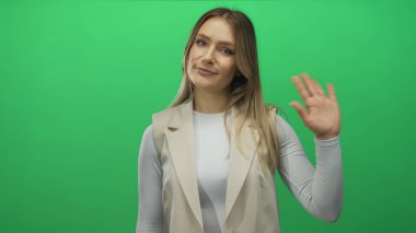 Yeşil arka plan duvarında gülümseyen bir kadın, beyaz bir gömlek ve bej bir yelek giyiyor, elini sallıyor, neşeli bir ifadeyle gündelik tarzını sergiliyor..