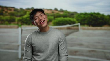 Tenis kortunda arka planda yeşillikle gülümseyen genç adam bulutlu bir gökyüzünün altında neşeli bir anı yakalıyor..