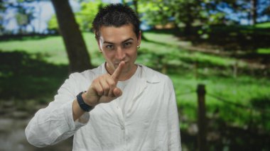 İspanyol adam, arkasında bulanık yeşil ağaçlar olan güneşli bir parkta boğazını keserek boyun hareketi yapıyor..