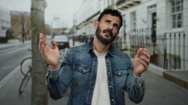 Kot ceketli sakallı bir adam sokakta bisiklet ve arabalarla duruyor, şehir ortamında el çırparken duygularını ifade ediyor..