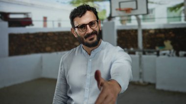Sakallı İspanyol bir adam, samimi bir hoş geldin jesti olarak açık hava basketbol sahasında gözlüklü ve gömlekli el sıkışma teklif ediyor..