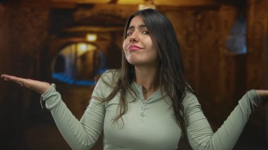 Sokak arasında genç bir İspanyol kadın, kollarını kaldırarak kafa karışıklığını dile getiriyor, günlük giysiler giyiyor, kentsel bir ortamda sıcak bir ışık altında..