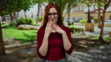 Kızıl saçlı kadın güneşli bir günde parkta dikilirken doğal yeşillik ve arka planda binalar olan günlük kıyafetlerini ve gözlüklerini sergiliyor..