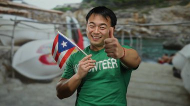 Deniz kenarında Puerto Riko bayrağı tutan genç bir adam baş parmağını kaldırmış, gönüllü gömleği giymiş, açık gökyüzü altında neşeli bir ifadeyle plaj manzarasının tadını çıkarıyor..