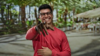 Güneşli bir terasta, hareketli bir açık hava restoranında gülümseyen genç bir adam, canlı bir şehirde mutluluk ve rahatlama yayıyor..