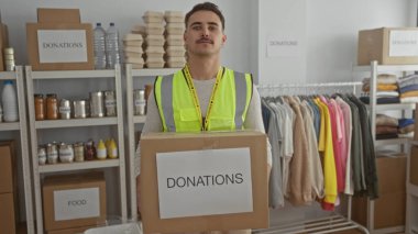 Yansıtıcı yelek giyen genç adam bağış kutusunu bağış odasında tutuyor. Etrafı giysi rafları ve konserve yiyeceklerle çevrili..