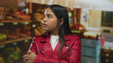 Kırmızı ceketli kadın meyva tezgahlarıyla açık pazarda duruyor, düşünceli ve kendinden emin görünüyor..