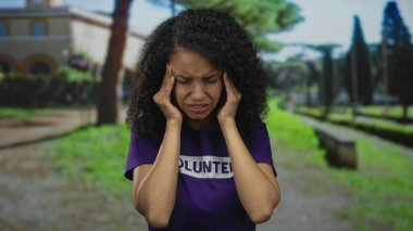 Gönüllü gömleğiyle parkta hayal kırıklığına uğramış bir kadın, yeşilliklerle çevrili, yol ve ağaçlarla çevrili açık bir ortamda duygularını ifade ediyor..