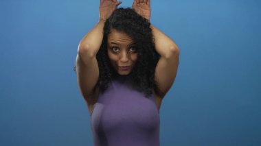 Çıplak omuzlu genç kadın, izole edilmiş mavi stüdyoda eli kulaklı tavşan kulağı yapar; neşe ve eğlence enerjisi..