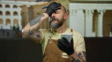 Sakallı ve dövmeli, şehir ortamında eldiven ve tulum giymiş, açık havada notlara odaklanmış düşünceli bir yüz ifadesi olan, konsantre olmuş bir adam..