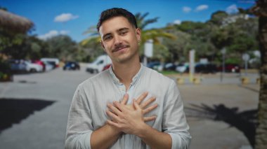 Açık mavi gökyüzünün altında park halindeki arabalar ve ağaçlarla çevrili güneşli bir şehir caddesinde kalbi olan genç İspanyol bir adam gülümsüyor..