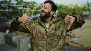 Sakallı, kamuflaj üniformalı bir asker yeşil bir parkta arka planda ağaçlarla dikilirken ve dışarıda karışık duygular gösterirken baş parmağıyla işaret veriyor..