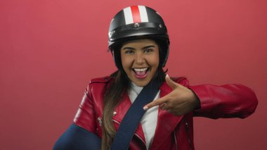 Kasklı gülümseyen kadın koluna işaret ediyor canlı kırmızı bir duvara yaslanıyor ve neşeli ruhunu vurguluyor.