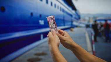 Deniz kıyısındaki büyük mavi bir geminin yanında Filipinli pesoları tutan adam, denizcilik veya seyahat bağlamında mali yatırım veya alım satım öneriyor..