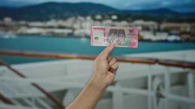 Deniz manzaralı bir yolcu gemisinde el ele tutuşan Endonezya rupiah banknotu, beyaz bir adamın kolu ve bulutlu bir gökyüzünün altındaki şehir silüeti..