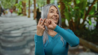 Gri saçlı yaşlı bir kadın, açık havada canlı bir jest yapıyor. Yeşillik ve arka planda insanlar var..