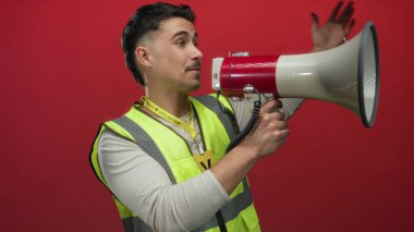 Yansıtıcı yelek giyen genç bir adam büyük bir tutkuyla kırmızı duvara karşı megafon kullanıyor. Gönüllülük ve iletişim ruhunu somutlaştırıyor..