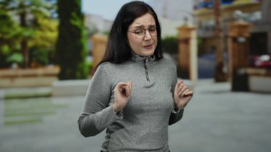 Esmer saçlı bir kadın, gözlüklü ve gri kazaklı bir sokakta dikiliyor. Ağaçlı ve kentsel geçmişli belirsiz bir ifadeyi canlandırıyor..