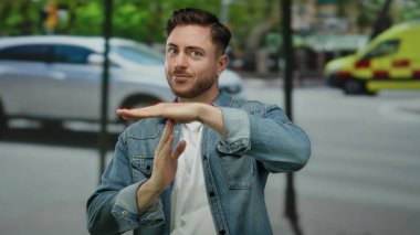 Sakallı genç adam şehir sokaklarında el hareketi yapıyor ve şehir ortamında gündüz vakti arka planda arabalar bulanıklaşıyor.